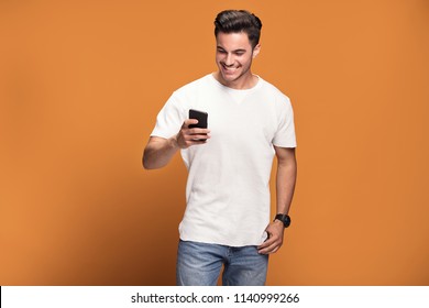 Photo Of Happy Handsome Man Holding Mobile Phone In Hand, Texting. Young Guy With Big Toothy Smile Posing On Yellow Background Wearing White T-shirt.