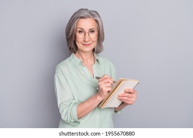 Photo of happy lovely pretty smiling mature woman author in glasses writing in notebook isolated on grey color background