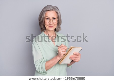Photo of happy lovely pretty smiling mature woman author in glasses writing in notebook isolated on grey color background