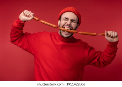 Photo of happy man with beard in glasses and red clothing. Holds and eats sausages, isolated over red background