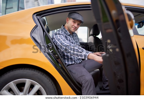 Photo of happy man in cap and\
plaid shirt looking to camera sitting in back seat in yellow\
taxi