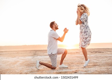 Photo of happy man making proposal to his excited woman with ring in gift box while walking on sunny beach