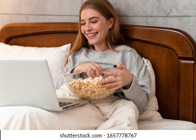 Photo Of A Happy Optimistic Pretty Young Woman Indoors At Home In Bed Using Laptop Computer Watch Film Eat Popcorn.