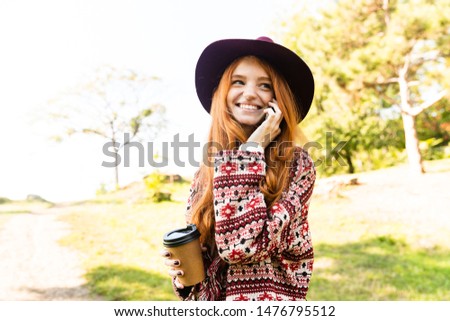 Photo of a happy pleased cutie young student redhead girl in autumn park talking by mobile phone.