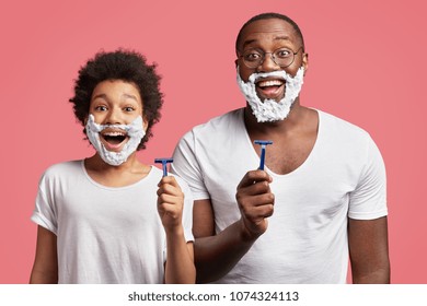 Photo of happy schoolboy with Afro hairdo attempts to shave like his dad, applies shaving gel on face and takes razor, wants to become older, stands next to father, enjoy free time together.