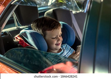 Photo Of A Happy Smiling Young Boy Riding In Car,using Social Network On Smartphone. Photo Of Young Male Passenger Sitting On Car Back Seat Using A Mobile. Toddler Sitting In A Car Playing With Phone.