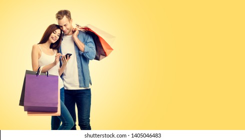 Photo Of Happy Smiling Young Couple With Shopping Bags, And Smartphone, Over Yellow Color Backround. Copy Space For Some Slogan Or Advertising Text. Caucasian Models - Online Shopping Concept.