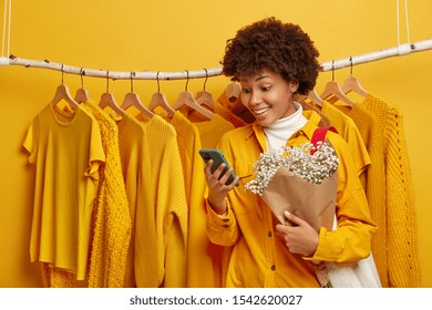 Photo Of Happy Woman Dresses For First Date, Stands Near Clothing Rack, Gets Pleasant Sms On Smartphone, Returns From Shopping Mall In Good Mood, Bought New Outfit On Sale, Holds Lovely Bouquet