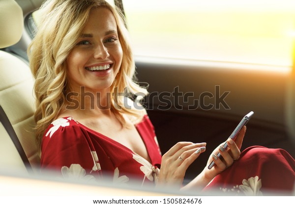 Photo of happy woman with phone in her hands\
sitting in back seat of car on\
summer.