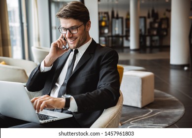 Photo Of Happy Young Businessman Wearing Formal Black Suit Using Laptop Computer And Talking On Smartphone In Hotel Hall