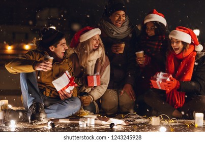 Photo Of A Happy Young Group Of Friends Sitting Outdoors In Evening In Christmas Hats Drinking Coffee.