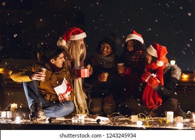 Photo Of A Happy Young Group Of Friends Sitting Outdoors In Evening In Christmas Hats Drinking Coffee.