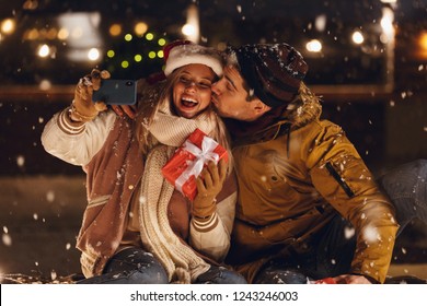 Photo of a happy young loving couple sitting outdoors in evening in christmas hat holding gift box take a selfie by phone. ภาพถ่ายสต็อก