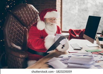 Photo Of Hard-working Grey Hair Santa Claus Prepare X-mas Christmas Sit Table Write Clip Board Organize Wish List Letters Order Wear Cap Headwear In Advent Spirit Lights House Indoors