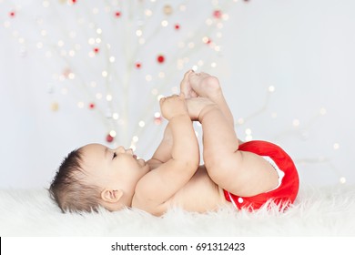 Photo Of A Healthy, Chubby Baby Lying On His Back On A White Sheepskin, Playing With His Toes, Wearing A Red Cloth Diaper/nappy With A Lit, White Christmas Tree In The Background.
