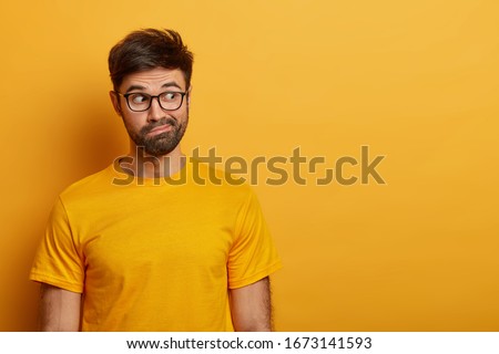 Photo of hesitant bearded man looks aside, smirks face and has puzzled expression, tries to decide something, dressed in casual yellow t shirt, poses over vibrant studio wall, wonders what he sees