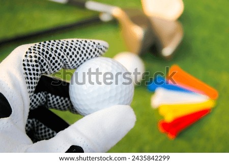 Photo of holding a golf ball with a golf glove on the left hand. The background has other equipment. and green grass.
