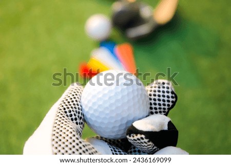 Photo of holding a golf ball with a golf glove on the left hand. The background has other equipment. and green grass.