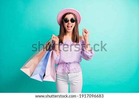 Photo of hooray brunette girl hand fist hold bags wear purple shirt cap trousers spectacles isolated on blue color background