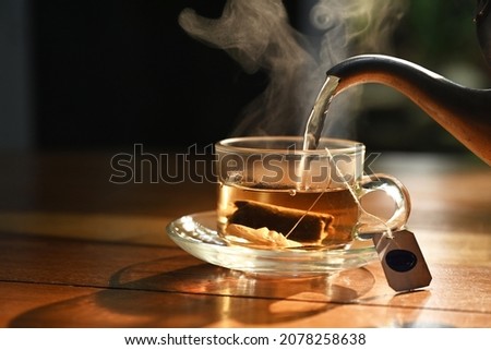 photo of hot water pouring into transparent cup with tea bag inside on wood table in morning sunlight