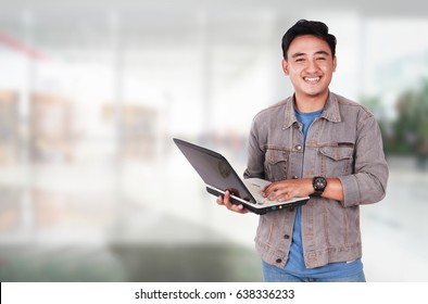 Photo image portrait of a cute young Asian male student standing and smiling while holding laptop and typing on it