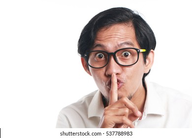 Photo image portrait of a funny young Asian businessman wearing glasses showing ssh sign, close up portrait with one finger on lips asking to silence gesture, over white background