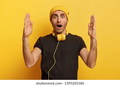 Photo Of Impressed Caucasian Man Makes Big Sign With Both Hands, Shapes Quite Huge Object, Impressed By Size, Keeps Mouth Widely Opened, Dressed In Black And Yellow Clothing, Uses Headphones