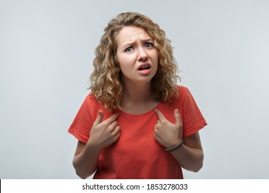 Photo of indignant blonde girl with curly hair in casual t shirt pointing fingers at herself, has dissatisfied expression. Studio shot, white background. Facial expression concept