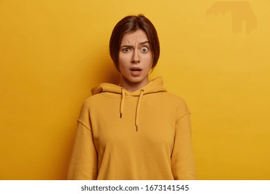Photo of indignant young European woman raises eyebrows, has unexpected expression, smirks face, wears casual hoodie, expresses wonder, poses against yellow background. Human face expressions concept
