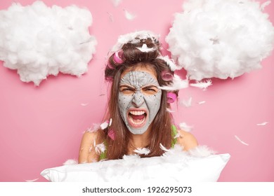 Photo Of Irritated Woman Has Bad Mood After Sleep Interruption Shouts Angrily At Someone Stands Under Flying Feathers Holds Pillow Applies Hair Rollers Facial Beauty Mask. Bad Morning Concept