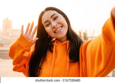 Photo Of Joyful Beautiful Asian Woman Smiling And Waving Hand While Taking Selfie Photo Outdoors