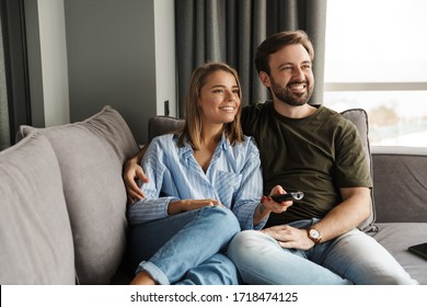Photo of joyful beautiful nice couple watching TV and using remote control while sitting on sofa at home