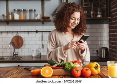 Photo of joyful caucasian woman holding smartphone while cooking salad with fresh vegetables in kitchen interior at home - Powered by Shutterstock