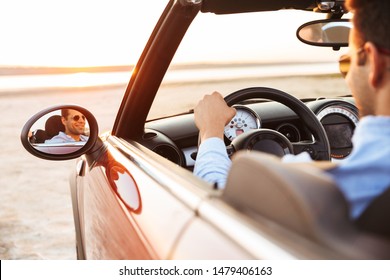 Photo Of Joyful Handsome Man Wearing Sunglasses Driving Convertible Car By Seaside At Sunrise