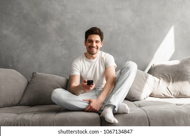 Photo Of Joyful Man 30s In Casual Clothing Sitting On Sofa In Living Room And Looking On Camera With Remote Control In Hand