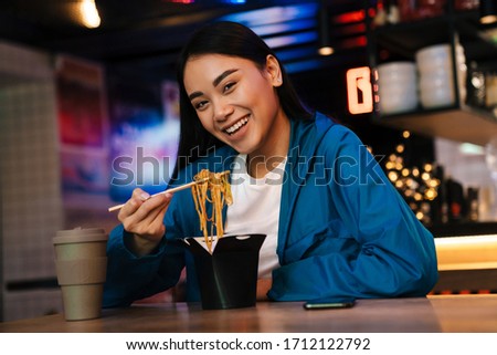 Photo of joyful nice asian woman eating chinese noodles and smiling while sitting in cafe