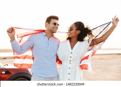 Photo of joyous multiethnic couple man and woman smiling and holding american flag while standing by car outdoors - Powered by Shutterstock