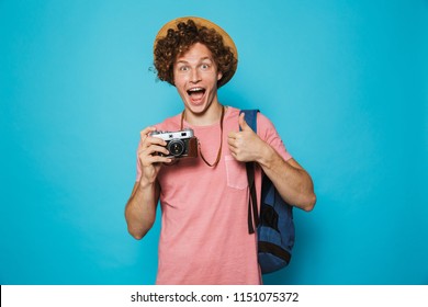 Photo Of Joyous Traveler Boy 18-20 With Curly Hair Wearing Backpack And Straw Hat Photographing On Retro Camera Isolated Over Blue Background