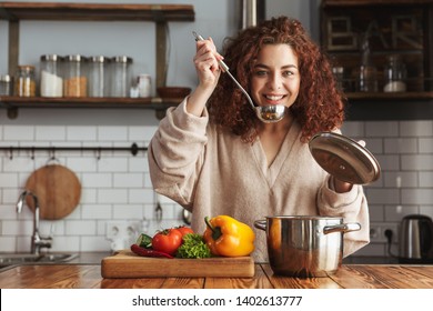 Photo Of Kind Caucasian Woman Holding Cooking Ladle Spoon While Eating Soup With Fresh Vegetables In Kitchen At Home