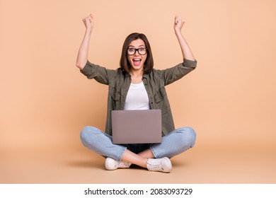 Photo of lady sit floor hold laptop rejoice raise fists wear glasses khaki shirt isolated beige color background