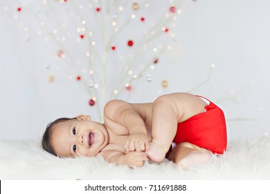 Photo Of A Laughing, Healthy, Chubby Baby Lying On His Side On A White Sheepskin, Playing With His Toes, Wearing A Red Cloth Diaper/nappy With A Lit, White Christmas Tree In The Background.