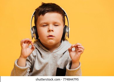 Photo of little concentrated boy child sitting on floor isolated over yellow background with eyes closed listening music with headphones.