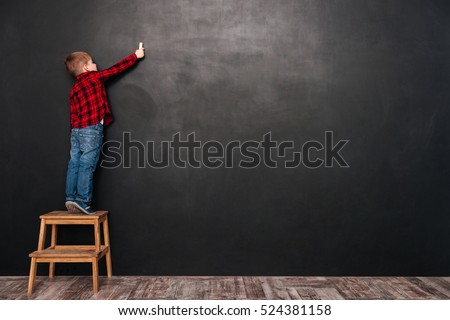 Photo of a little cute child standing on stool near blackboard and drawing on it.