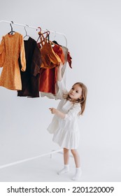 Photo of a little girl who chooses what clothes to wear among those offered on the hanger. Photo on a white studio background