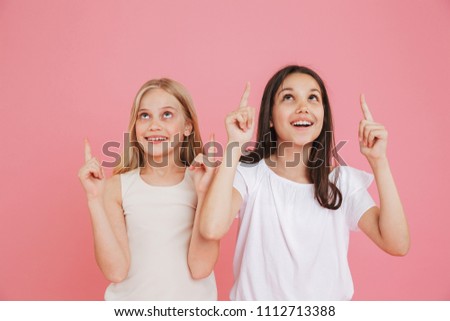 Photo of little girls 8-10 years old wearing casual clothing pointing fingers upward at copyspace while looking up together isolated over pink background