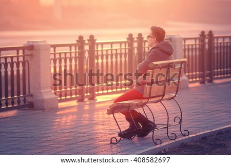 Photo of Lonely Man Sitting on a Bench during Sunset. Young Man Sitting on Wooden Bench. Sunset Male Outdoor Background