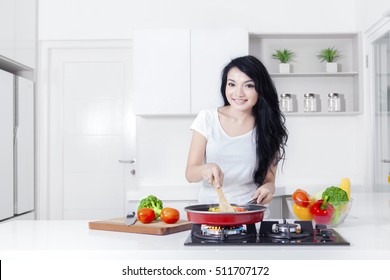 Photo of a lovely Asian woman smiling at the camera while cooking vegetable with a frying pan in the kitchen