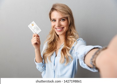 Photo of lovely blond businesswoman smiling and holding credit card while taking selfie isolated over gray background - Powered by Shutterstock