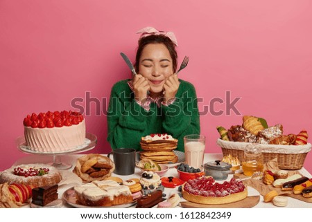 Photo of lovely millennial girl holds knife and fork, thinks what to eat first, sits at big table full of cookies, pastries and cakes, eats different desserts during holidays, has bad eating habits