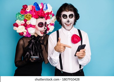Photo of lovely spooky couple man lady hold direct finger telephone find undead friend social network wear black dress death costume roses headband suspenders isolated blue color background